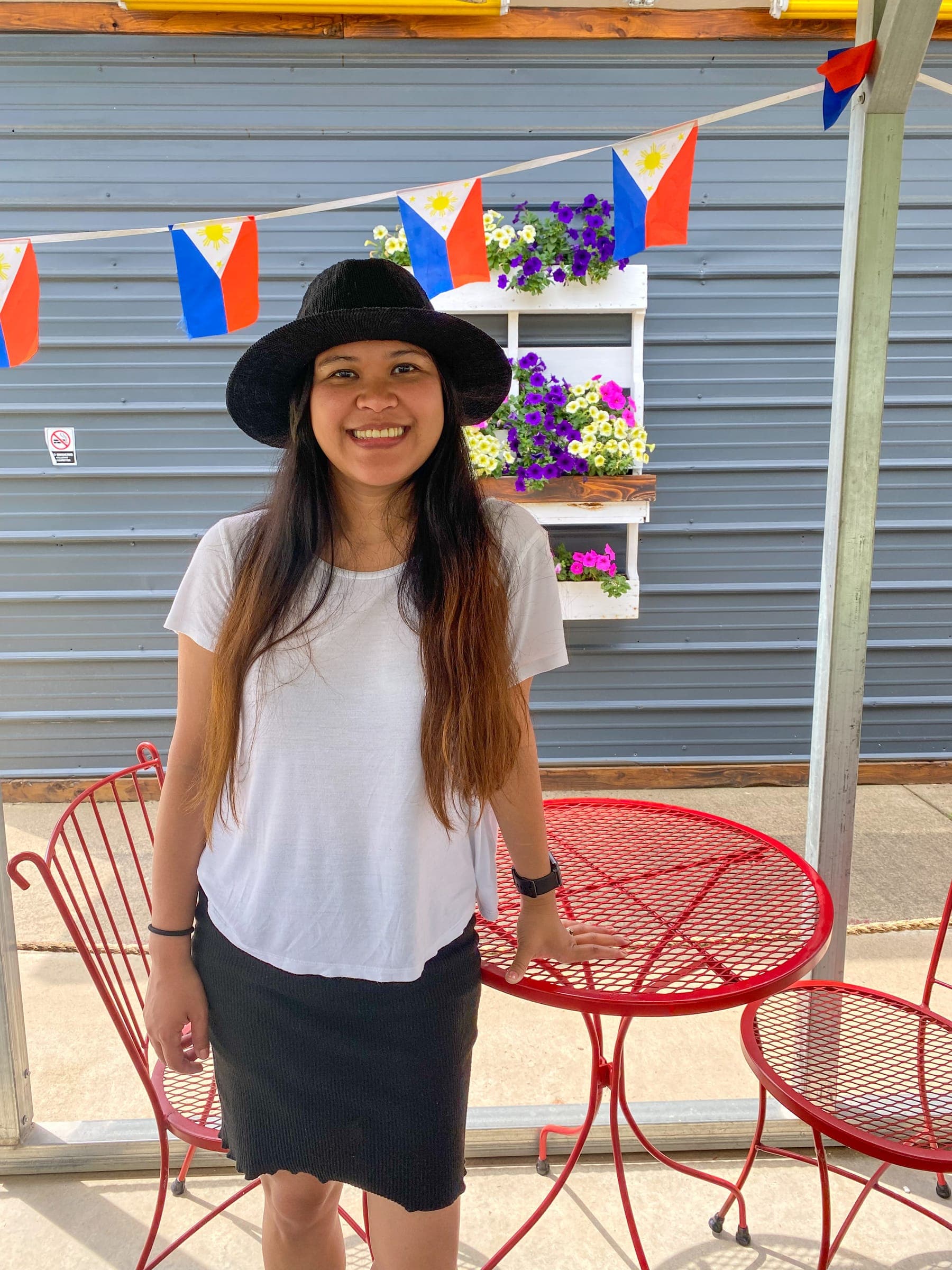Kristy smiling in a black hat, standing by a red bistro table with Philippine flag bunting behind her