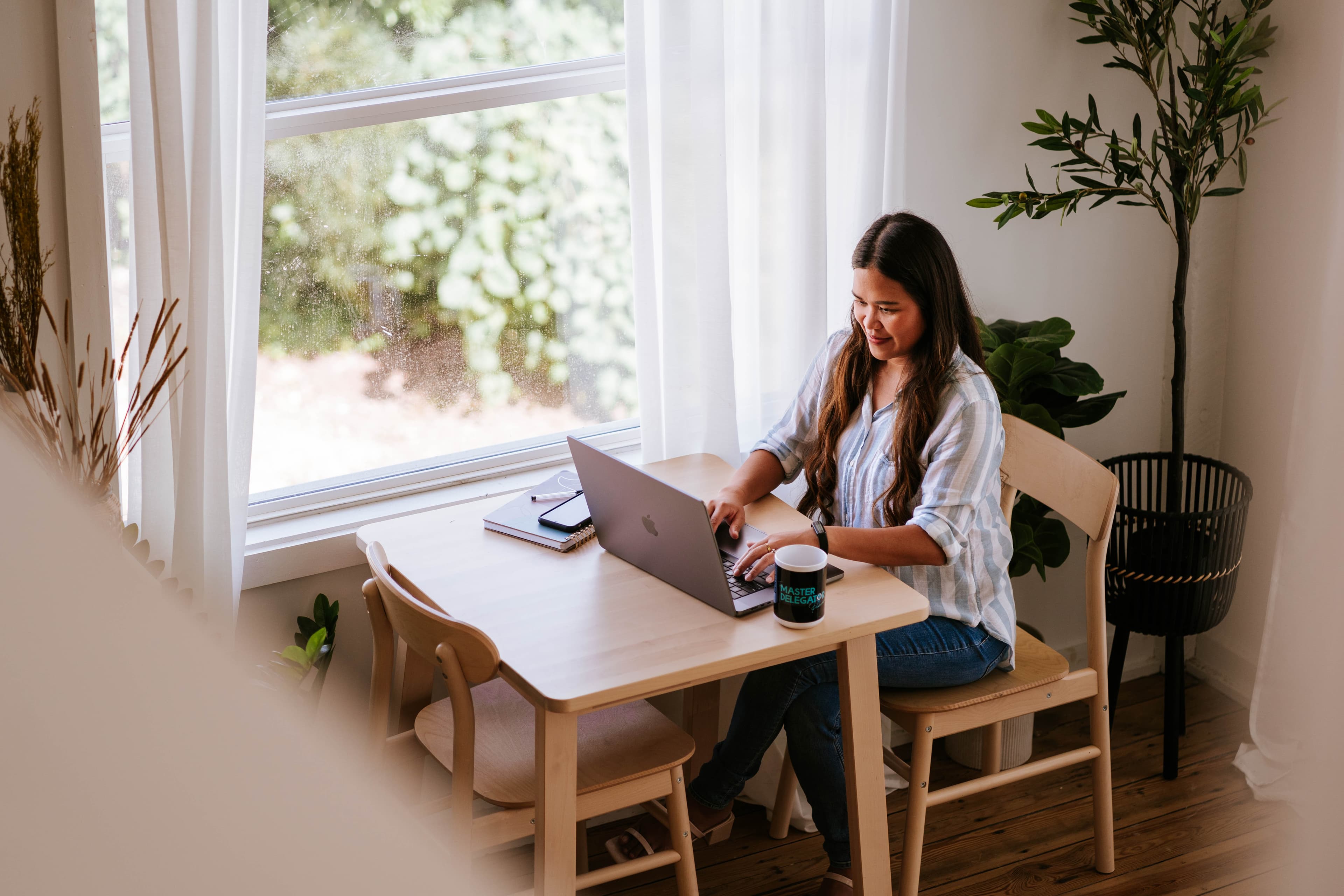 Kristy Yoder, business coach for agency owners, working on her laptop at a sunlit wooden table beside a window